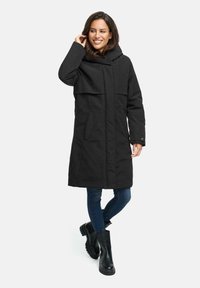 Zwarte, knielange parka met een capuchon, voorzien van een dubbele laag aan de voorkant, zijzakken en een soepele textuur. Gecombineerd met blauwe jeans en zwarte laarzen.