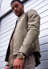Veste bomber en daim vert olive avec des zips, superposée à une chemise brune à boutons, assortie à une montre dorée et un pantalon gris.