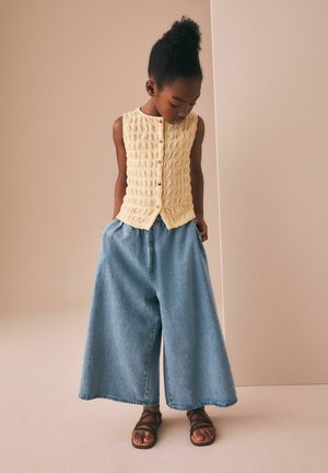 Dieťa s tmavou pokožkou a kučeravými vlasmi stojí a pozerá sa dole, oblečené v bezrukávom béžovom pletenom topu, širokých modrých džínsových nohaviciach a hnedých sandáloch.