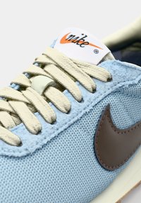 Jasnoniebieski sneaker z teksturowanej tkaniny, kremowymi sznurowadłami i brązowym logo Nike w kształcie łyżwy po boku. Posiada wyściełany język i kontrastową podeszwę.