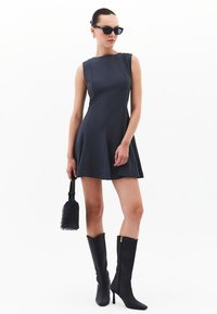 Vestido sin mangas de color marino con un cuerpo ajustado y falda acampanada. Combinado con botas negras hasta la rodilla y un pequeño bolso de mano negro tejido.