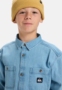 Niño que lleva un gorro de punto amarillo y una camisa de botones de mezclilla azul claro con dos bolsillos en el pecho, uno de los cuales muestra un parche del logo de Quiksilver.