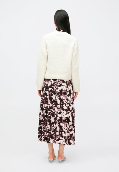 Biely sveter s textúrou a dlhými rukávmi, skombinovaný s kvetinovo potlačenou plisovanou maxi sukňou v odtieňoch fialovej, čiernej a krémovej. Viditeľné sú podpätky.
