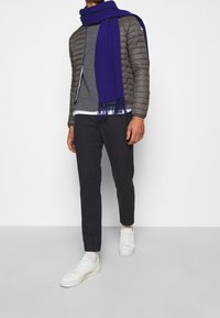 Grijze gewatteerde jas, gestructureerde grijze trui, blauwe sjaal, donkere broek en witte sneakers. De sjaal heeft franje randen. Eenvoudige, casual outfit.