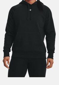 Sweat à capuche noir avec poche avant, capuche à cordon de serrage, et poignets côtelés. Présente un petit logo sur la poitrine. Le tissu semble doux et extensible.