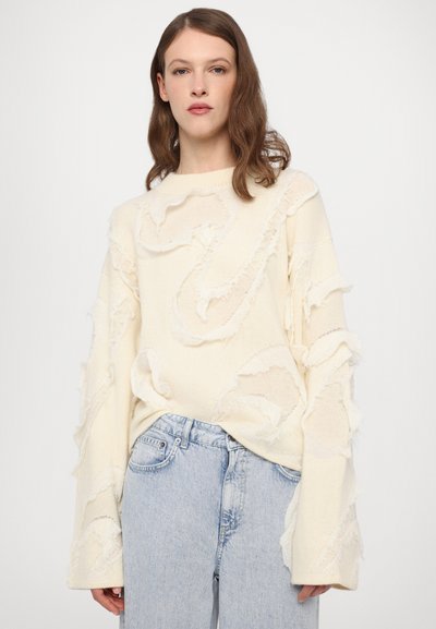 Cream sweater with textured, frayed accents and long sleeves, paired with light blue high-waisted jeans. Soft knit material, loose fit.