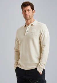 Polo-shirt beige à manches longues avec col, patte de boutonnage à trois boutons et logo brodé. Il présente une poche à rabat sur la manche gauche.