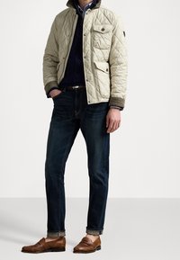 Quilted beige jacket with a collared neckline, button details, and front pockets, paired with dark blue jeans and brown loafers.