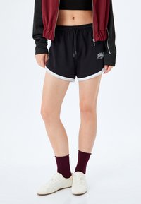 Czarne spodenki sportowe z białym wykończeniem, elastycznym pasem ze sznurkiem oraz małym logo. Połączone z białymi butami i burgundowymi skarpetami.