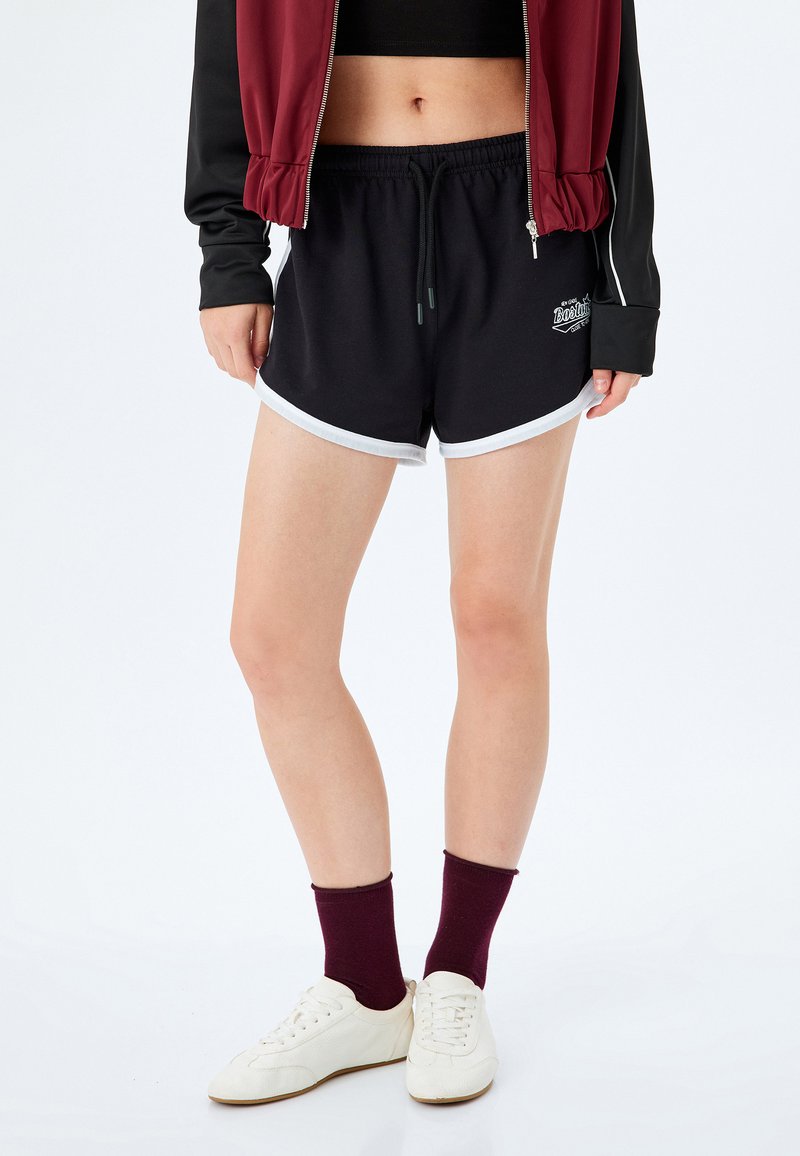 Czarne spodenki sportowe z białym wykończeniem, elastycznym pasem ze sznurkiem oraz małym logo. Połączone z białymi butami i burgundowymi skarpetami.