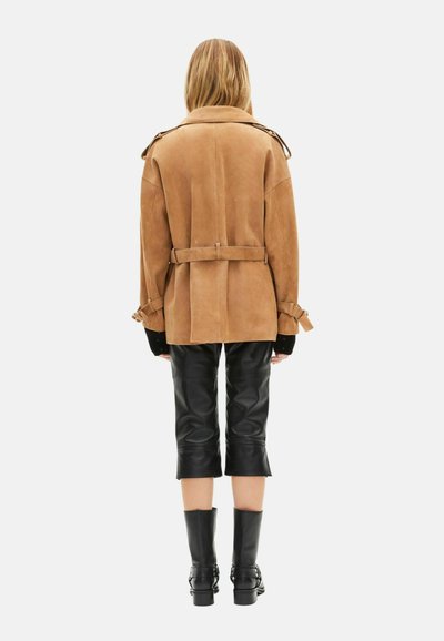 Chaqueta de ante marrón con cinturón, de ajuste amplio y mangas con puños, combinada con pantalones cortos de cuero negro y botas negras hasta la rodilla.