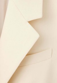 Gros plan sur le revers crème d'un blazer et une poche passepoilée avec une texture de tissu lisse.