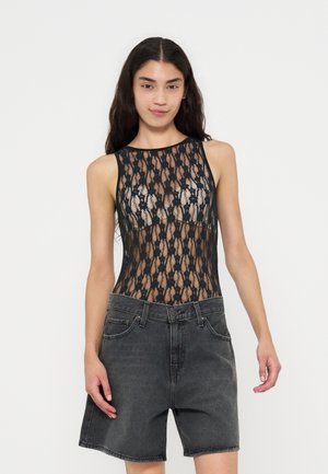 Person med langt mørkt hår bærer en sort ærmeløs blondetop over en beige bh og løstsiddende sorte denimshorts med høj talje, står neutralt.