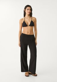 Crni bikini top s valovitim rubovima, uparen s labavim crnim hlačama širokih nogavica. Model nosi smeđe sandale s otvorenim prstima. Minimalistički dizajn, mekani materijal.