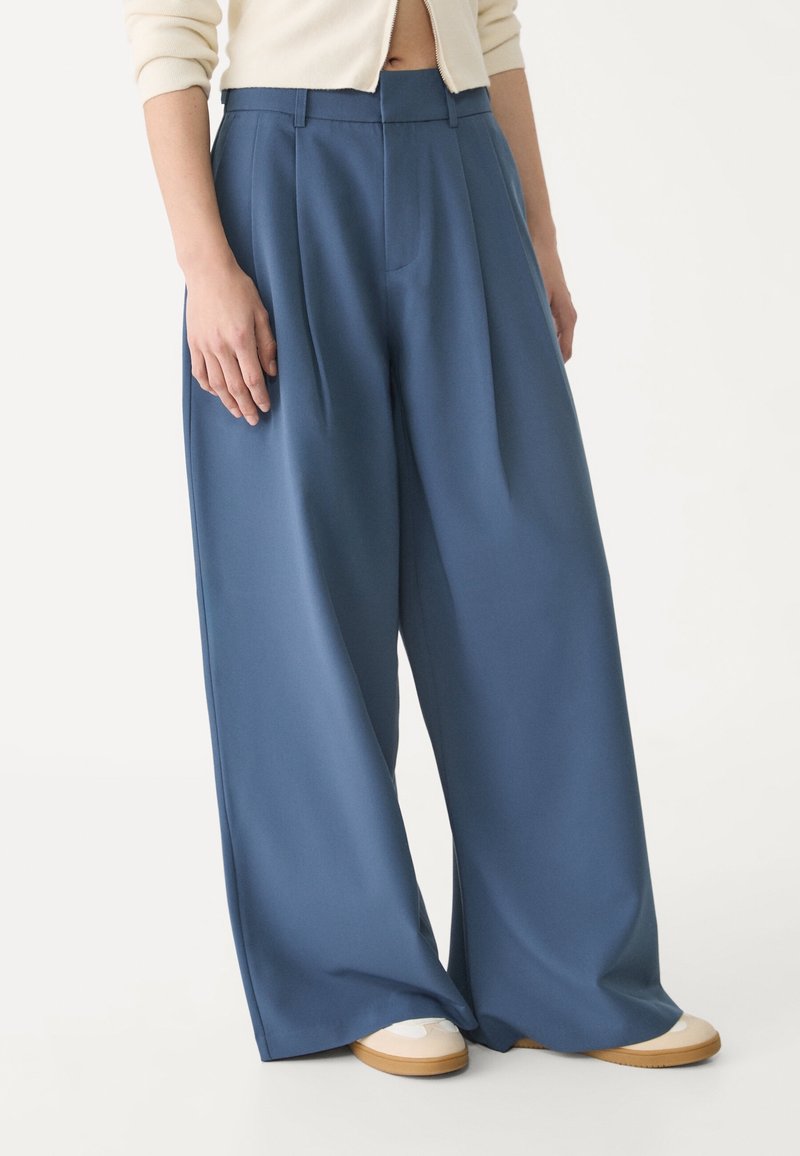 Personne portant un pantalon large bleu ample et des baskets beiges, debout devant un fond blanc uni.