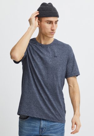 Mann trägt eine dunkelgraue Strickmütze, ein blau meliertes T-Shirt und blaue Jeans, zieht die Mütze zurecht, während er zur Seite schaut.