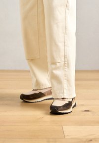 Pieds portant des baskets beiges et brunes avec des semelles épaisses, posés sur un sol en bois clair, avec un pantalon beige couvrant les chevilles.