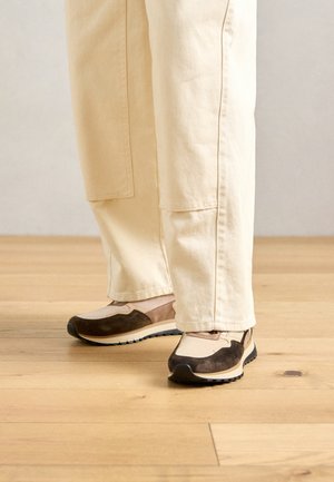 Voeten in beige en bruine sneakers met dikke zolen, staand op een lichte houten vloer, met beige broek tot aan de enkels.