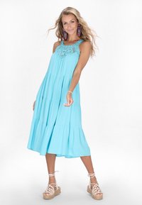 O rochie fără mâneci, cu volane, de culoare albastră deschisă, cu un decolteu din dantelă. Asortată cu sandale espadrille cu bretele de culoare bej. Țesătură netedă și fluidă.