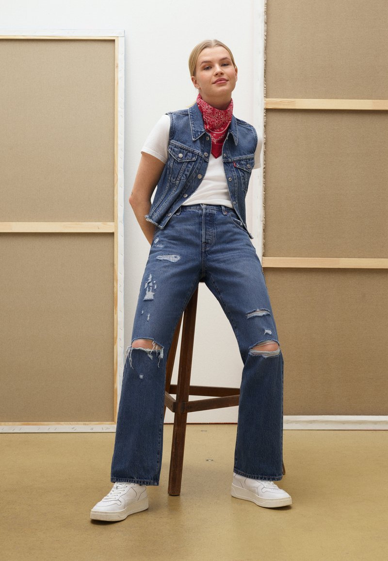 Chaleco de mezclilla sobre una camiseta blanca, combinado con jeans azules rasgados y zapatillas blancas. Una bandana roja añade contraste. Taburete de madera clara al fondo.