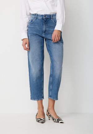 Woman wearing cropped blue jeans, white buttoned shirt, and black-and-white zebra print high heels, standing against a white background.