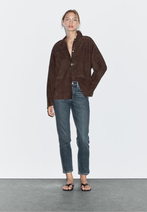 Brun mockaskjorta med krage och framknäppning, kombinerad med mörkblå jeans och svarta sandaler. Enfärgad bakgrund.