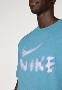 Turkizna kratka majica s kratkimi rokavi iz mehkega materiala, ki ima �čez prsa velik valovit roza Nike logo.