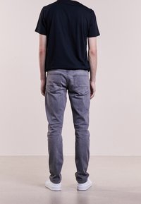 Persona de pie de espaldas, vistiendo una camiseta negra, jeans grises y zapatillas blancas sobre un fondo neutro.