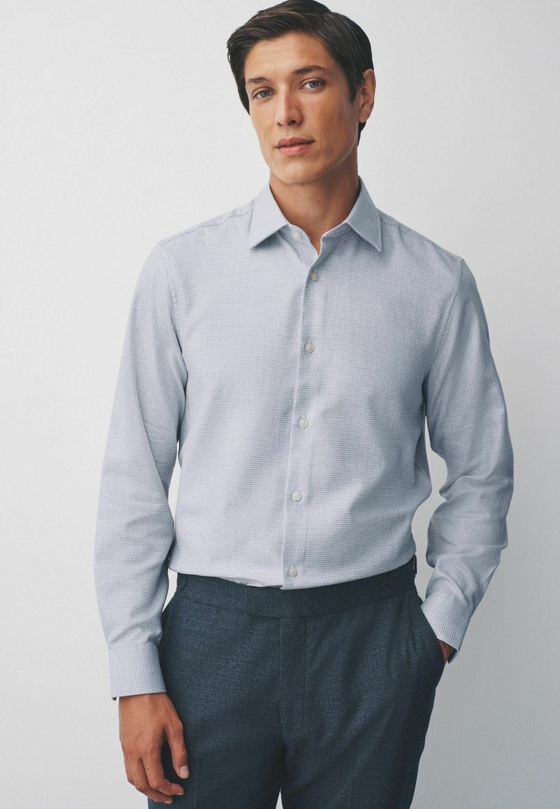 Light blue patterned button-up shirt with a collar and long sleeves, paired with dark trousers. Features a subtle texture and white buttons.