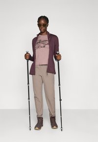 Donna con occhiali da sole, che tiene bastoni da trekking neri, vestita con giacca malva, t-shirt con grafica, pantaloni beige e stivali da trekking marroni, in piedi di fronte a uno sfondo neutro.