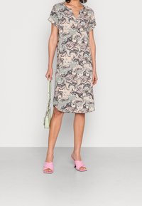 Mujer vestida con un vestido de patrón paisley en tonos pastel, sosteniendo un bolso verde claro y sandalias de tacón rosas sobre un fondo liso.