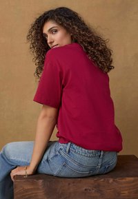 Vinröd bomulls t-shirt med korta ärmar, i kombination med ljusblå högmidjade jeans. Texturen verkar mjuk. Bakgrunden har en varm, neutral nyans.