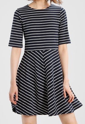 Mujer vistiendo un vestido azul marino con rayas blancas horizontales en la parte superior y rayas diagonales en la falda, de pie con los brazos relajados.