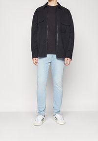 Chaqueta negra con cremallera, cuello corto, dos bolsillos delanteros, combinada con una camiseta negra y jeans ajustados de un azul claro, zapatillas blancas con acentos negros.