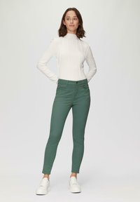Weißes Langarmshirt mit strukturierten Nähten, kombiniert mit grünen, enganliegenden Hosen und weißen Sneakers. Einfaches Design, ohne auffällige Muster oder Akzente.