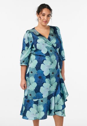 Frau mit lockigem Haar, die ein blau-grünes, geblümtes Wickelkleid mit dreiviertel Ärmeln trägt und vor einem weißen Hintergrund steht.