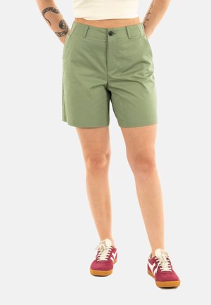 Pantaloni corti in cotone verde con taglio dritto, chiusura a bottone e due tasche laterali. Indossati con sneaker bordeaux e bianche.
