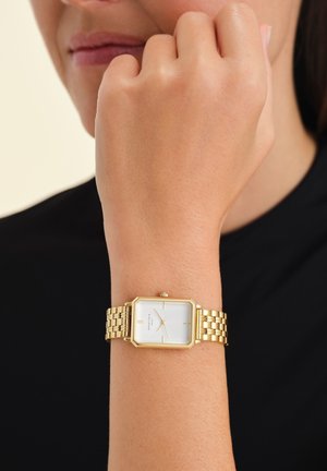 Montre octogonale dorée avec un cadran blanc, dotée d'un bracelet en maillons métal et de marqueurs d'heure noirs simples. Portée au poignet contre un tissu sombre.