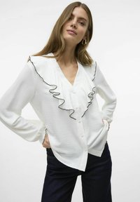 Blusa blanca con un ribete frontal rizado bordeado en negro. Presenta mangas largas y un escote en V, cierre de botones y tejido texturizado.