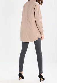 Chaqueta ligera beige con cuello alto y cinturilla elástica, combinada con jeans gris ajustados y tacones de aguja negros.
