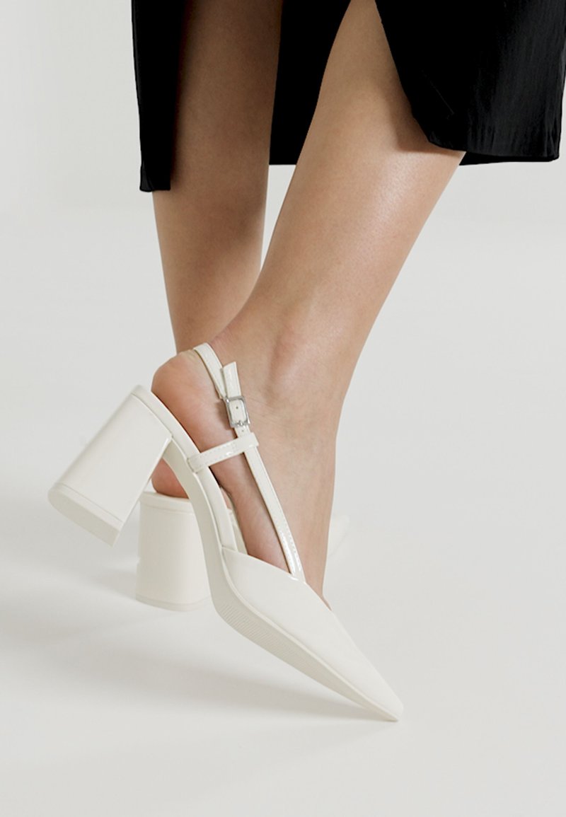 Escarpins blancs à talons ouverts avec un bout pointu, un large talon bloc et une bride ajustable à la cheville. Matériau lisse et brillant avec des accents fins.