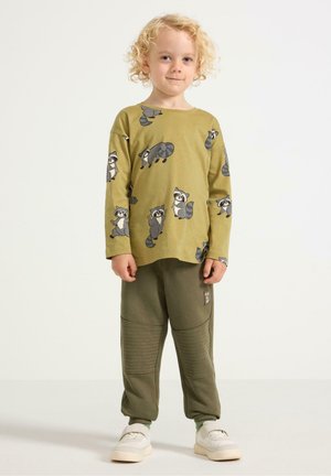 Olijfgroen overhemd met lange mouwen en wasbeer prints, gecombineerd met bijpassende olijfgroene joggers met textuurpanelen en een klein logo-opdruk. Witte schoenen.