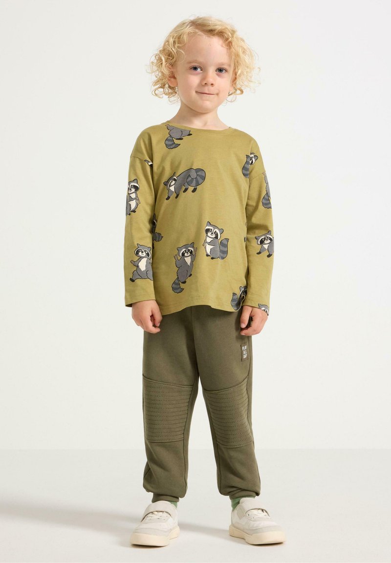 Oliven grøn langærmet skjorte med vaskebjørn tryk, parret med matchende oliven joggers med teksturerede paneler og et lille logo-patch. Hvide sko.