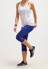 Kvinna som sträcker ut benet, klädd i en vit Nike-topp, blå tights, svarta Nike-sneakers och en svart aktivitetsklocka på handleden.