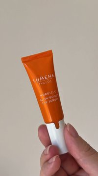 Oranger Tubus des Lumene Nordic-C Glow Boost Augenserums, weiße Düse, glatte Textur, bedrucktes Etikett. Hand hält den Tubus vor hellem Hintergrund.
