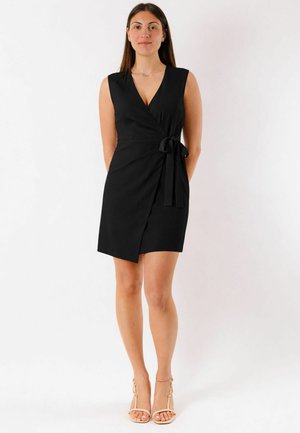 Donna in piedi a piedi nudi che indossa un abito nero avvolgente senza maniche, annodato in vita, su uno sfondo bianco uniforme.