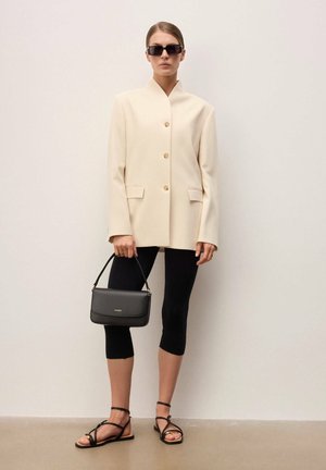 Naine kannab üle suureks kasvanud kreemikat bleiserit, musti lühikesi retuuse, musti sandaale ja päikeseprille, ning hoiab väikest musta käekotti ühetaustase seina taustal.