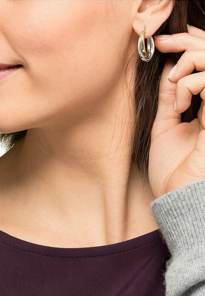 Sølv- og guld-hoops øreringe med et lagdelt design, hængende fra en cirkulær lås, båret af en person, der rører ved en med en sweater.