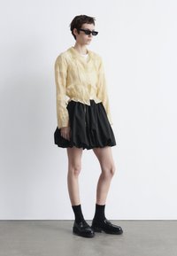 Yellow sheer button-up blouse with lace detail, black voluminous skirt, black loafers, and dark socks. Simple, modern design with layered textures.
