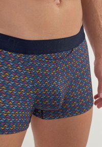 Shorts with a navy elastic waistband featuring "HOM" logo. The fabric displays a multicolored zigzag pattern on a dark background.